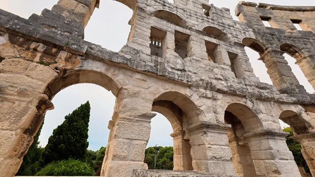 View from inside the Pula Arena showing the iconic limestone arches and stone tiers under a clear sky. This well-preserved Roman amphitheater showcases ancient masonry and classical architecture.
