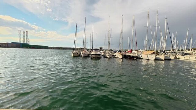 A wide shot of numerous sailboats and yachts moored in the calm waters of the Adriatic Sea at Pula. The background features industrial port structures and a dramatic sky with shifting clouds