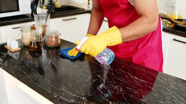 Arabian man wipes kitchen counter with cleaner and microfiber cloth removing grease for hygiene and food safety. Young guy keeps home apartment kitchen spotless feeling calm and proud after housework.