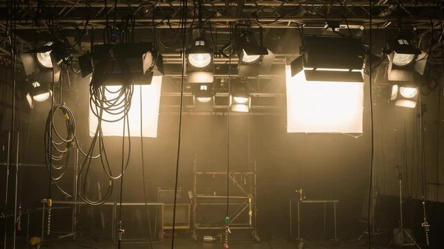 Medium shot of a television studio lighting grid featuring spotlights and softboxes casting a warm glow over an empty set with cables dangling from the ceiling rig.