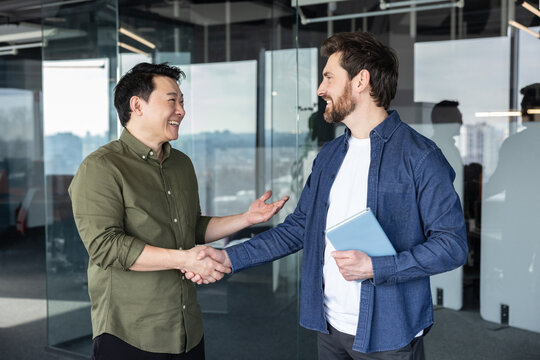 Two male businesspeople from different ethnicities happily shaking hands, building trust and cooperation during a successful negotiation in a modern glass office
