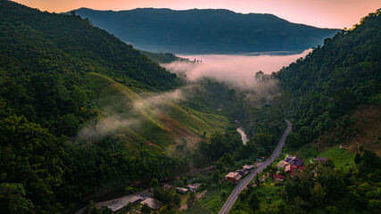 A breathtaking aerial view of Sapan Village in Nan, Thailand, covered by a magical sea of mist at sunrise. Warm morning light illuminates the mountain valley while fog drifts through lush green hills © Narong Niemhom