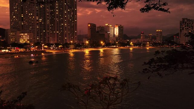 Night view from Hon Chong cape overlooking the north bay of Nha Trang, Vietnam. City lights and glowing windows along the coastline with dark sea and illuminated buildings.