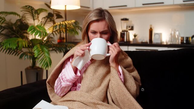 Adult woman at night home feels sick sore throat wrapped in blanket holds tea takes sip uses tissues. Caucasian girl on sofa at evening wipes nose checks temp rests breath drinks tea hopes for relief.