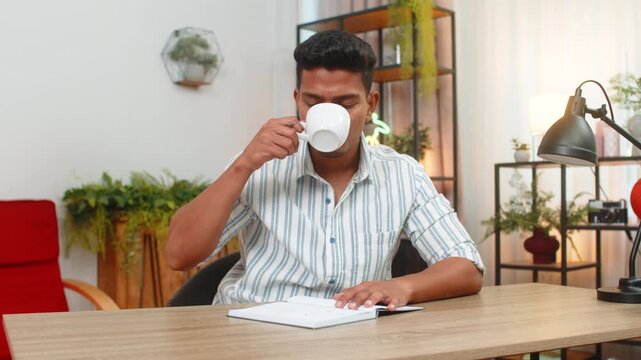 Indian man at home drinks coffee and writes goals in notebook ticks boxes and plans day with drive. Arabian guy at desk reviews list adds tasks sets priorities smiles feels focus and motivation aim