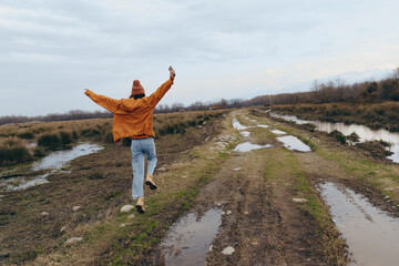 Fototapeta premium A woman lifestyle concept showing a joyful person in casual clothes walking on a muddy rural road with arms raised. The cloudy sky and natural landscape create a carefree outdoor mood.