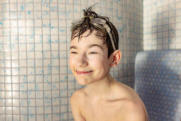 Young boy with wet hair and swimming goggles smiles in a tiled shower, water droplets visible on...