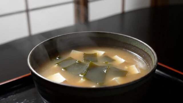 Close-up shot of a steaming bowl of miso soup with tofu on a black tray in a traditional Japanese setting with warm tones.