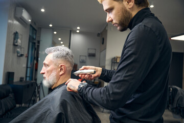 Obraz premium Hairdresser is cutting hair of handsome bearded mature man in salon