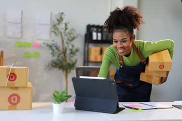 Smiling young woman engaging in a video call on a tablet while holding a package in her office.