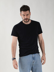 Portrait of a 35 years old bearded man in a basic black t-shirt and blue jeans. He looks directly at the camera with a hostile, tense, menacing and focused gaze.