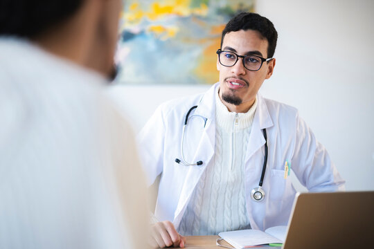 A doctor in a white coat and stethoscope consults with a patient, reviewing notes and a laptop in a modern office setting.