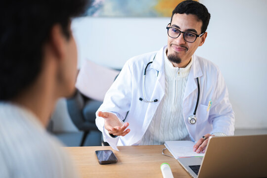A doctor in a white coat consults with a patient, gesturing while looking at notes and a laptop.