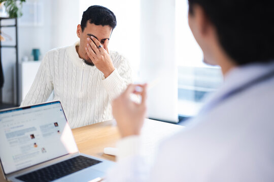A man rubs his eyes while speaking with a medical professional in an office setting, with a laptop open on the table.