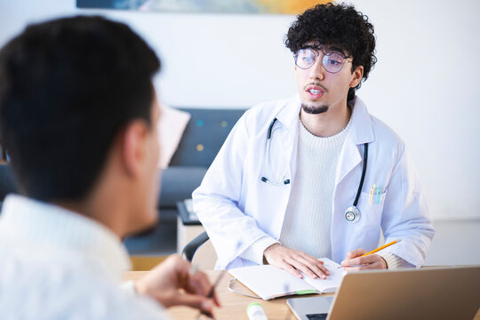 A doctor in a white coat with a stethoscope consults with a patient, taking notes in a journal and using a laptop.