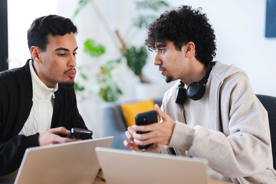Two young men collaborate on a project, discussing ideas while looking at their phones and laptops in a modern office setting.