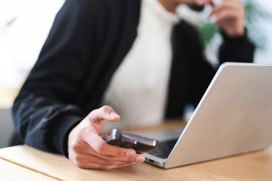 A person uses a mobile phone while working on a laptop at a wooden desk, demonstrating modern multitasking and remote work.