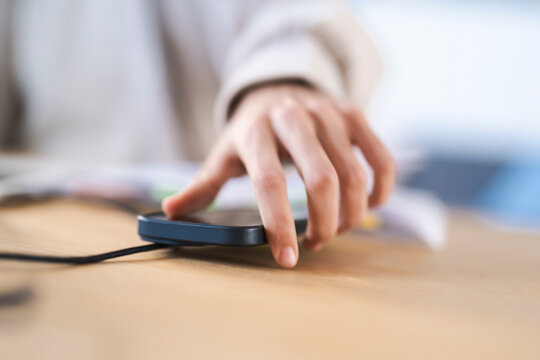 A person's hand reaches for a dark-colored mobile device connected to a charging cable on a wooden desk.