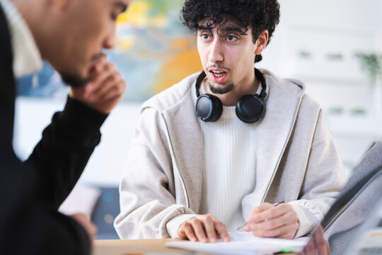 Two young men collaborate on a project, one speaking while the other listens intently, with headphones around his neck.