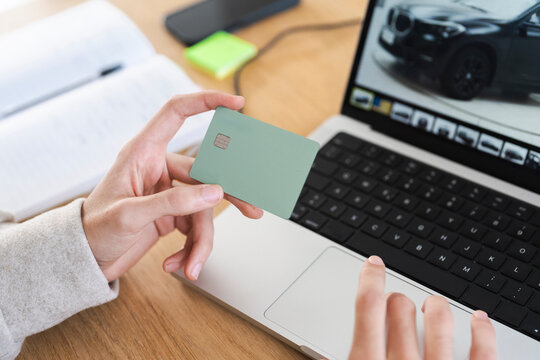 A person holds a payment card near a laptop, with a notebook and phone on the desk, suggesting online transactions or research.
