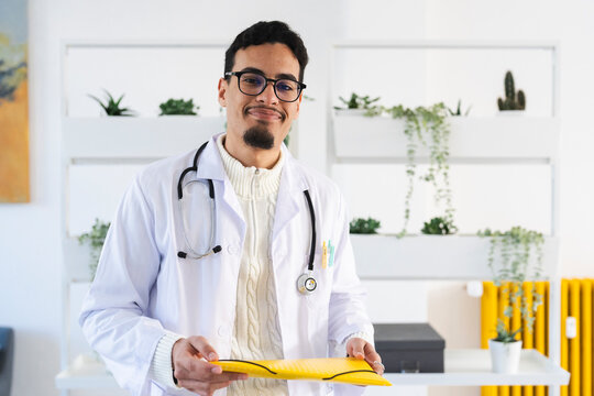 A smiling male doctor in a white coat and stethoscope holds a yellow folder. He is in a modern office with plants on shelves.