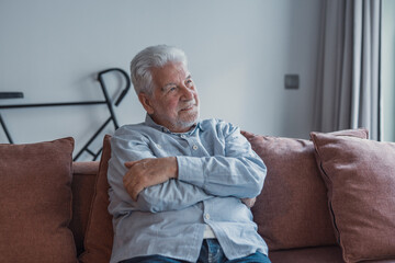 Fototapeta premium Elderly man sits on a sofa at home looking outside the window. He feels calm and reflective while enjoying a serene moment in his daily life.