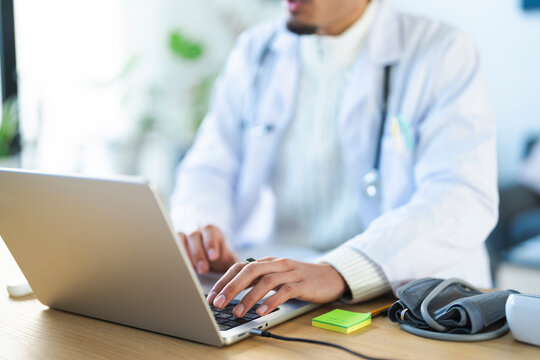 A medical professional in a white coat uses a laptop, with a stethoscope and blood pressure cuff nearby, indicating a focus on patient care and data management.