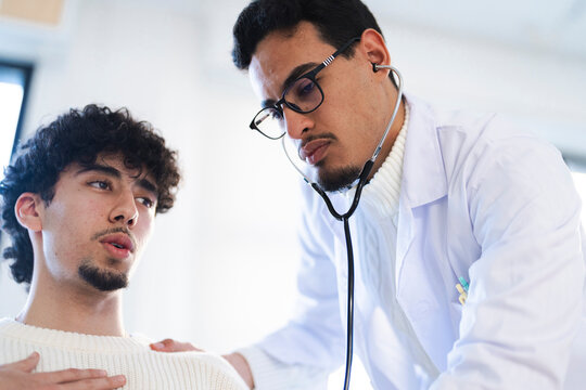A doctor wearing glasses and a white coat uses a stethoscope to examine a young man. The patient looks concerned during the medical checkup.