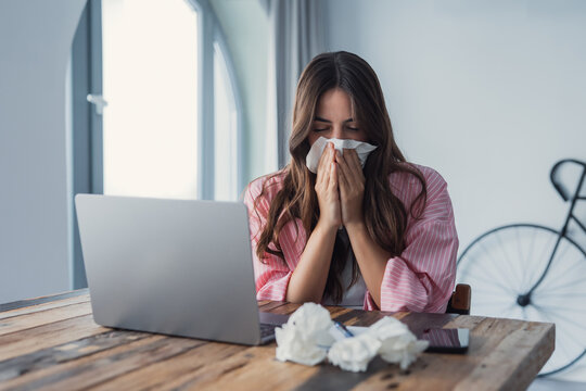 Frustrated young woman feeling sick at home, suffering from cold, flu, or fever, holding a tissue to her face while consulting a doctor online via video call, illustrating illness, discomfort