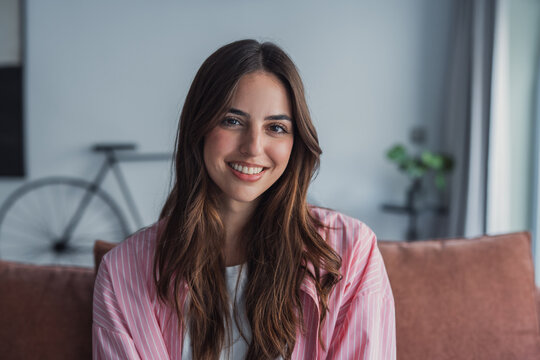 Happy attractive young adult woman housewife sitting on sofa looking at camera in modern cozy home, smiling single 20s lady beautiful face relaxing on couch alone enjoy wellbeing posing for portrait