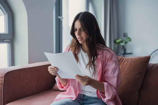 Worried young woman sitting at home reading a paper letter, looking stressed and confused after receiving negative financial news or an unexpected notice, illustrating concern and uncertainty