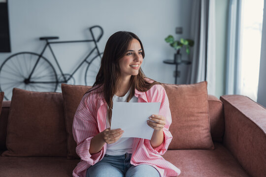 Smiling young woman sitting comfortably on a sofa at home while reading a letter with positive news, feeling relieved and happy after receiving an approval or favorable notice in an official document