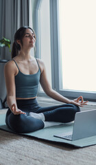Fototapeta premium Young woman practicing yoga and deep breathing on a mat at home, following an online tutorial on her laptop, calm and focused, wellness, mindfulness, fitness lifestyle indoors
