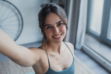 Fototapeta na wymiar Young woman sitting on yoga mat at home taking a first-person POV selfie with her smartphone, smiling at the camera while practicing yoga and showing a personal lifestyle moment