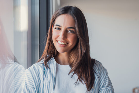Head shot portrait confident positive woman with healthy toothy smile looking at camera, standing near window, successful happy businesswoman entrepreneur student posing for profile picture