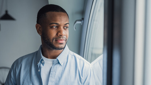 Thoughtful millennial African American man looking at window away in deep thoughts, thinking over problems, bad news, difficult tasks, feeling bored, sad. Portrait of pensive guy, homeowner