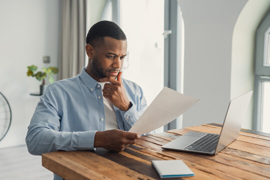 Concentrated African American manager reading letter, paper handout, analyzing company statistics, thinking about financial risks, comparing results. Thoughtful black worker considering business offer