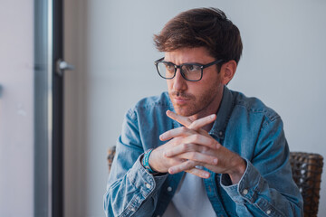 Thoughtful young businessman in stylish eyeglasses, looking out the window with a serious...