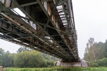 Fototapeta premium Wide angle perspective from below showing complex industrial steel girder network of railway bridge spanning river toward stone supports and forested shoreline under overcast sky.