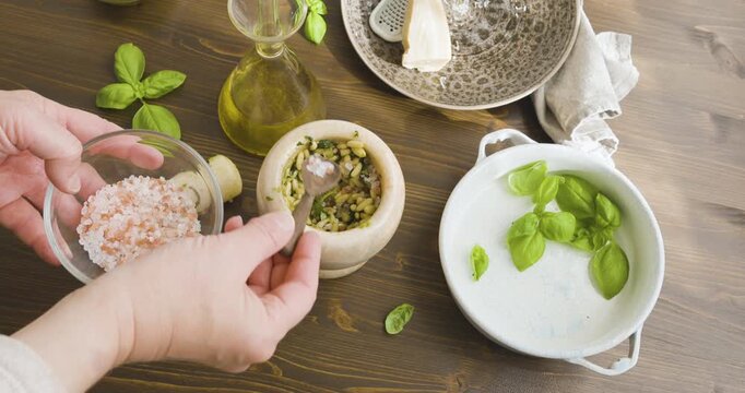 Person add pink sea salt to marble mortar while making traditional Genovese pesto. Close up of hands using small wooden spoon to add Himalayan salt into mortar with pine nuts and basil. Olive oil and