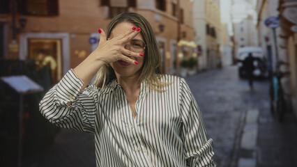 Obraz premium Woman hides face with hand wearing striped blouse and glasses on narrow cobblestone street in european city; playfulness.