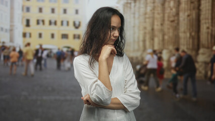 Fototapeta premium Woman in white dress hand on chin on crowded street near cathedral columns and old building; quiet contemplation.