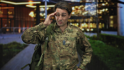 Obraz premium Man soldier in camouflage with backpack and dog tags, hand on forehead outside a lit building; duty fatigue.