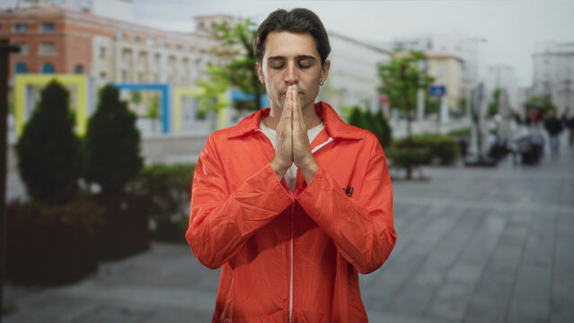 Man in orange jumpsuit presses bare hands together in prayer, eyes closed, standing on a city street in front of buildings and planters; remorse.