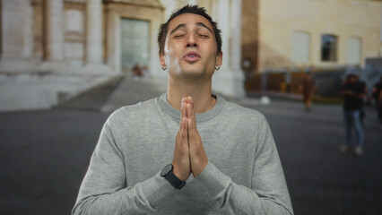Naklejka premium Young man in old town outdoors, gesturing with hands in prayer near historical architecture, conveying hope and peace.