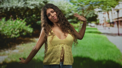 Fototapeta premium Young woman with hand to hair and arm out, wearing yellow knit top showing midriff on street; carefree joy.