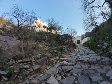 A historic white-painted main gate and stone pathway leading to a religious shrine situated on a rugged hill within the Aravalli Range in Nuh district