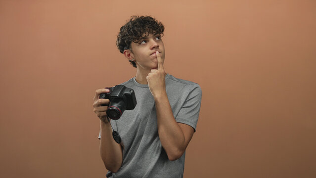 Young man holding dslr camera with finger to lips for silence against warm brown studio backdrop, gazing upward; thoughtful curiosity.