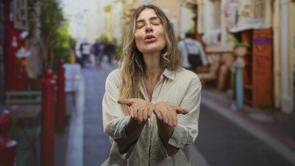 Fototapeta premium Woman covers her face with hands then opens them to blow a kiss toward camera on a lively street; playful joy.
