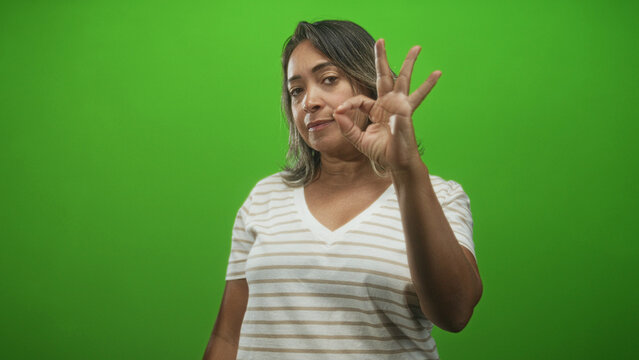 Hispanic woman wearing striped t shirt shows ok hand gesture with fingers near camera in green screen studio; confidence approval.